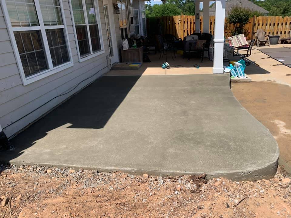 Curved concrete patio slab poured behind a home, tying into an existing wood fence line