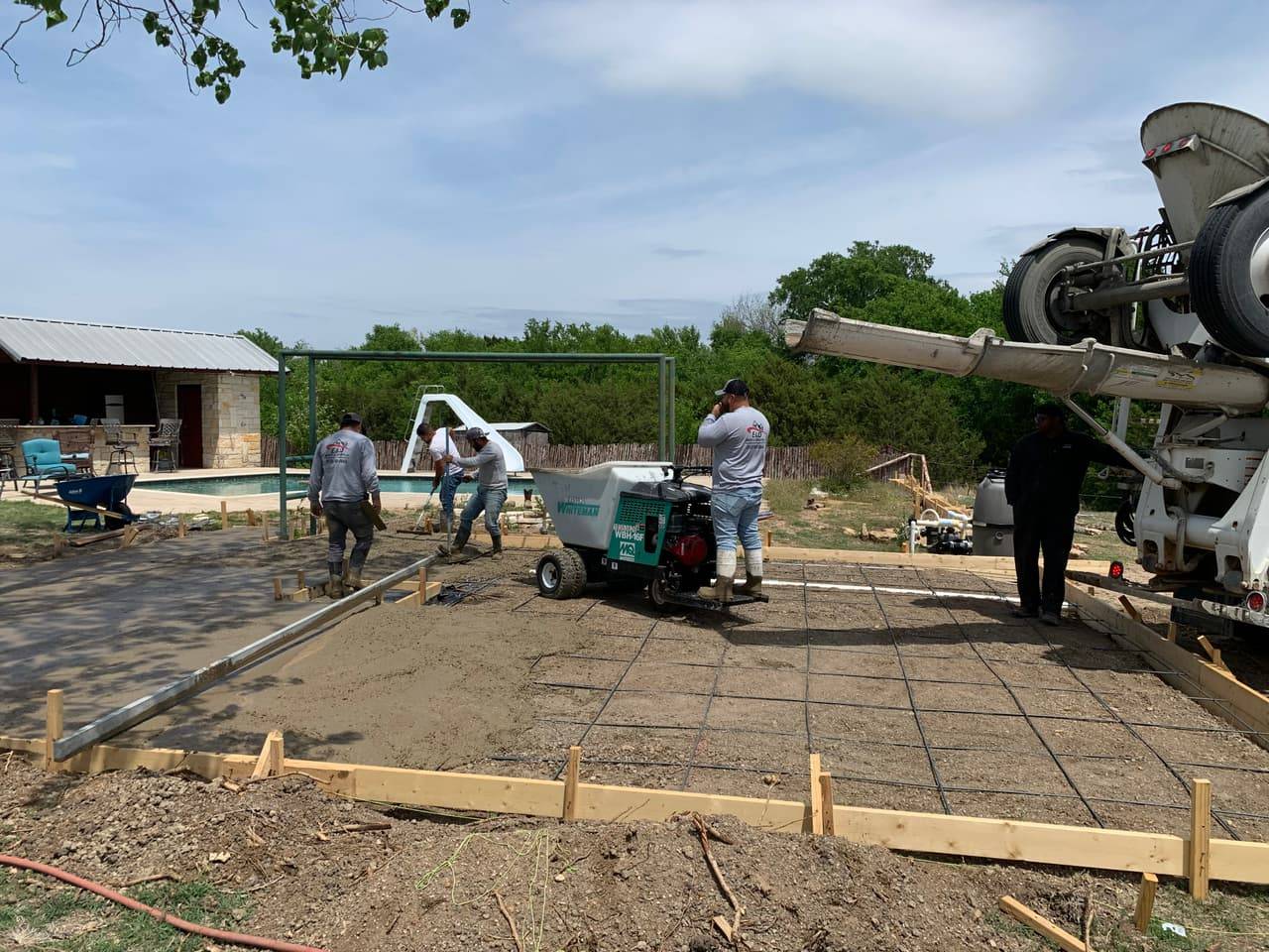 Crew pouring and finishing a concrete patio slab beside an in-ground pool